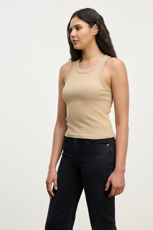 A woman with long dark hair wears the SARI TANK TOP by Velvet by Graham & Spencer in beige, paired with black jeans, standing against a plain light background.