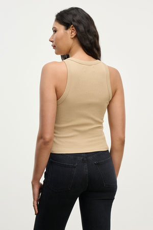 A woman faces away in the SARI TANK TOP by Velvet by Graham & Spencer—a beige ribbed essential paired with black jeans, set against a plain light background.