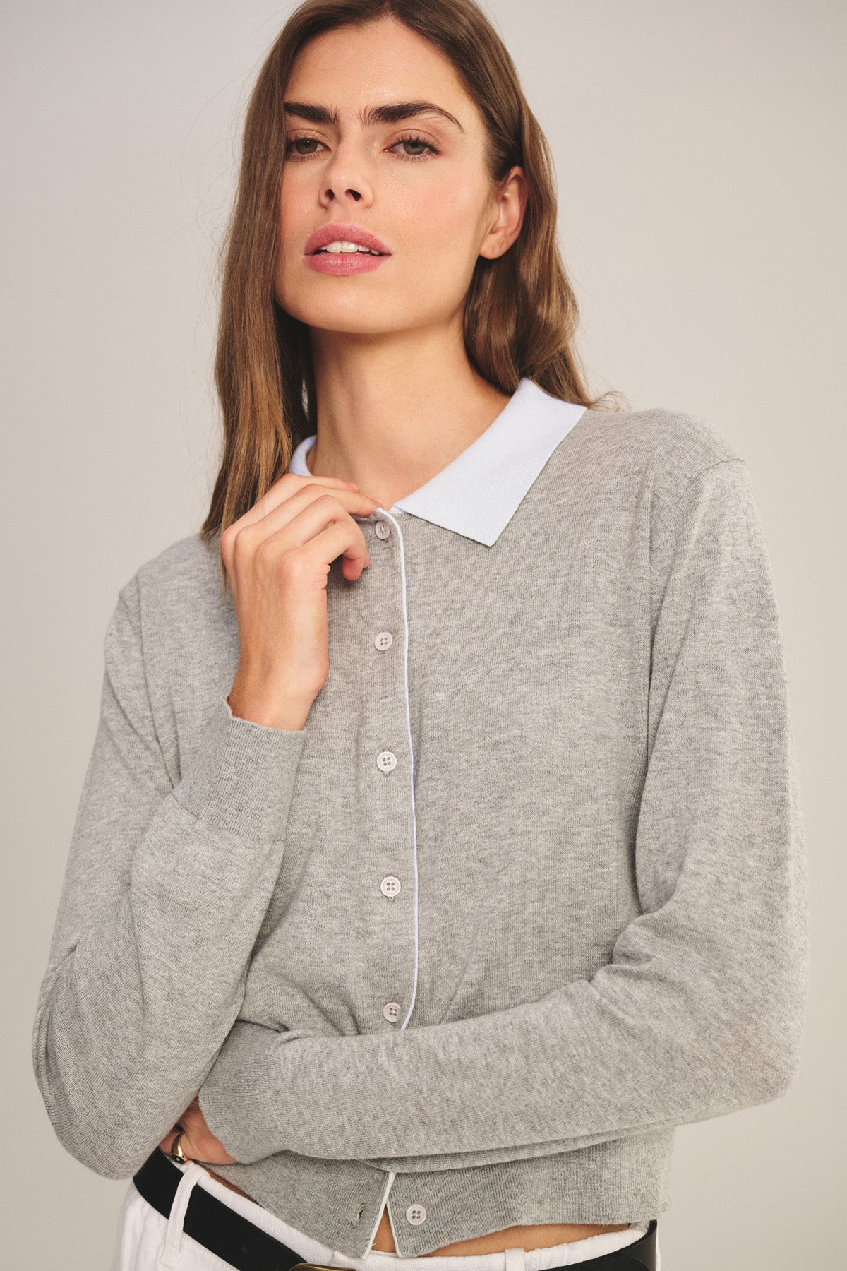 A woman with long brown hair wears the Velvet by Graham & Spencer RIVEN CARDIGAN—a light gray cotton-cashmere cropped cardigan—over a light blue collared shirt, standing against a plain background.-41277142696129