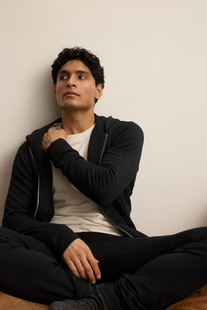 A man with short, curly dark hair sits cross-legged on the floor, wearing the Velvet by Graham & Spencer RODAN HOODIE over a white shirt and black pants, looking slightly to the side.
