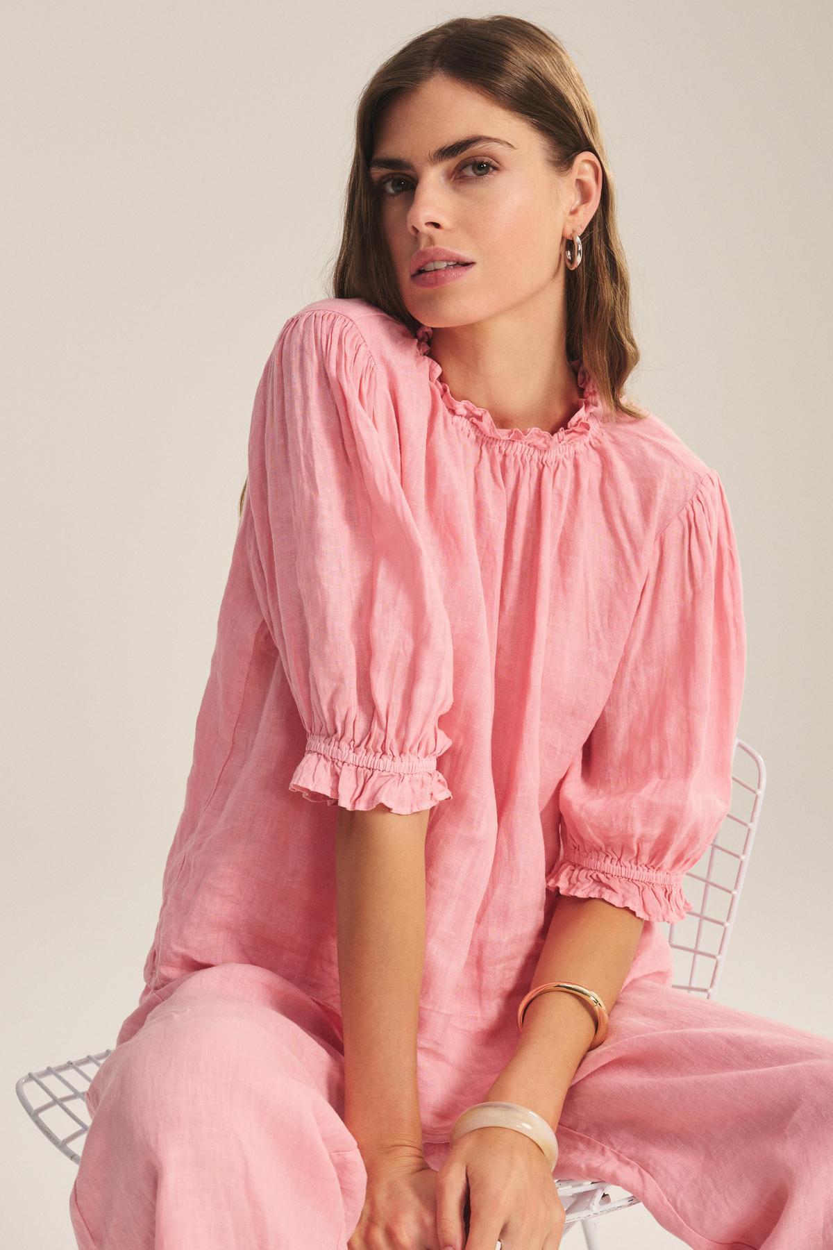A person with long hair sits on a white wire chair, wearing the ROSABEL LINEN TOP by Velvet by Graham & Spencer, paired with matching pants, hoop earrings, and two bangles.-41296981852353