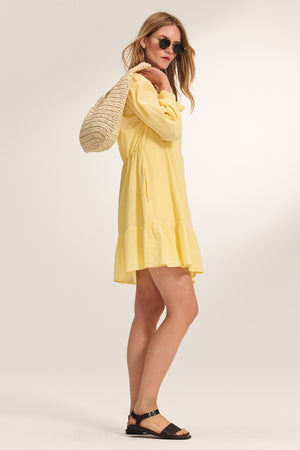 A woman models the SAMANTHA DRESS by Velvet by Graham & Spencer, a yellow ruffled dress with a smocked waist, styled with black sandals, sunglasses, and a woven bag against a neutral background.