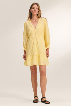 A woman wears the SAMANTHA DRESS by Velvet by Graham & Spencer, a light yellow cotton voile long-sleeve button-down with a smocked waist and ruffled hem, paired with black sandals against a neutral background.
