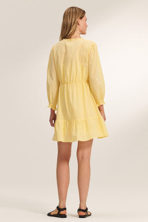 A woman faces away, wearing the SAMANTHA DRESS by Velvet by Graham & Spencer—a light yellow cotton voile knee-length dress with a smocked waist and ruffled hem—paired with black sandals against a neutral background.
