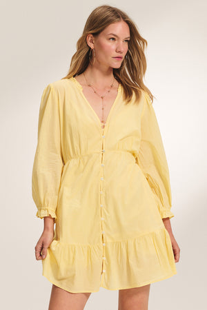 A woman poses against a plain background wearing the SAMANTHA DRESS by Velvet by Graham & Spencer—a light yellow, long-sleeve, lightweight cotton voile dress with a V-neckline and ruffled hem.