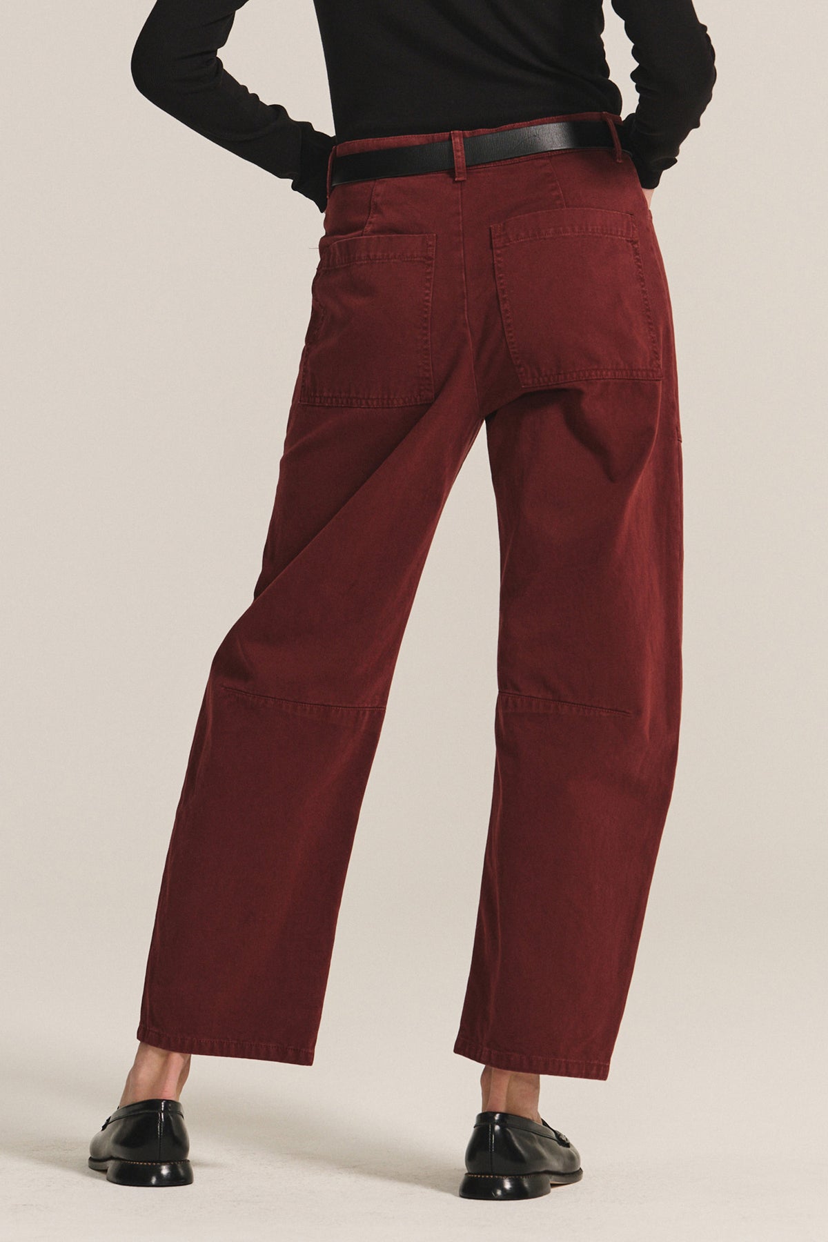 Person wears Velvet by Graham & Spencer's BRYLIE SANDED TWILL UTILITY PANT—high-waisted, loose maroon trousers with patch pockets—paired with a black belt, black long-sleeve top, and black shoes, shown from the back.-40235015012545