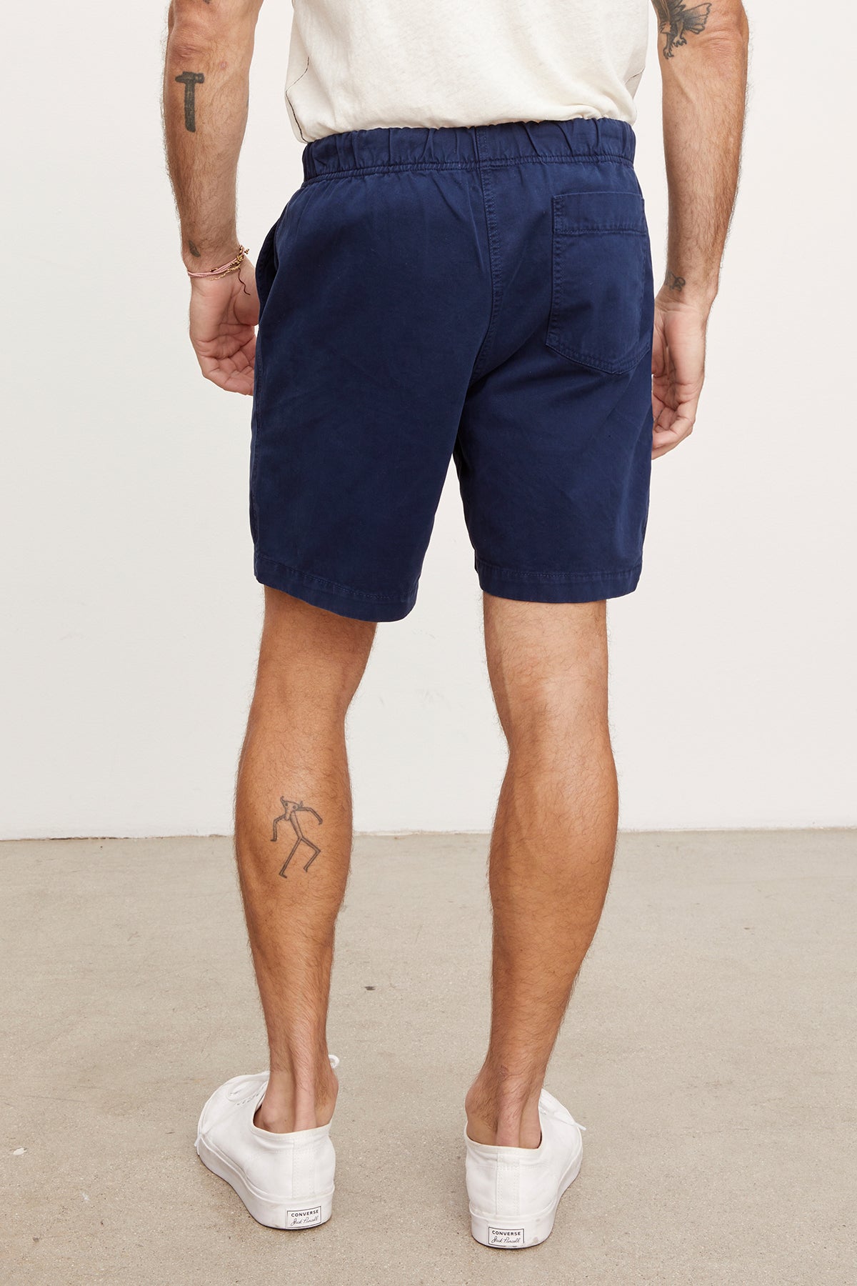 A man viewed from behind wearing navy blue cotton twill Velvet by Graham & Spencer Fielder shorts and white sneakers, standing with hands in pockets; visible tattoos on his legs.