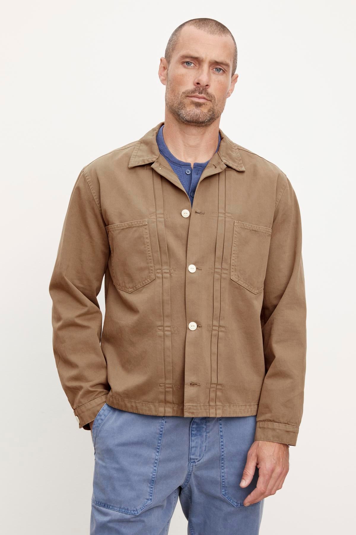 A man in a Velvet by Graham & Spencer FLANNERY SANDED TWILL BUTTON-UP JACKET and blue jeans standing against a plain background, exuding a vintage vibe.-36388077633729