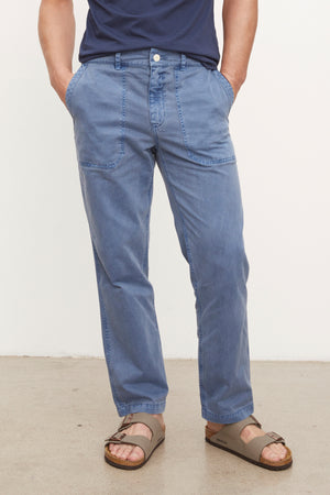 A man in a blue shirt and TOBY SANDED TWILL PANT from Velvet by Graham & Spencer is standing in front of a white wall.
