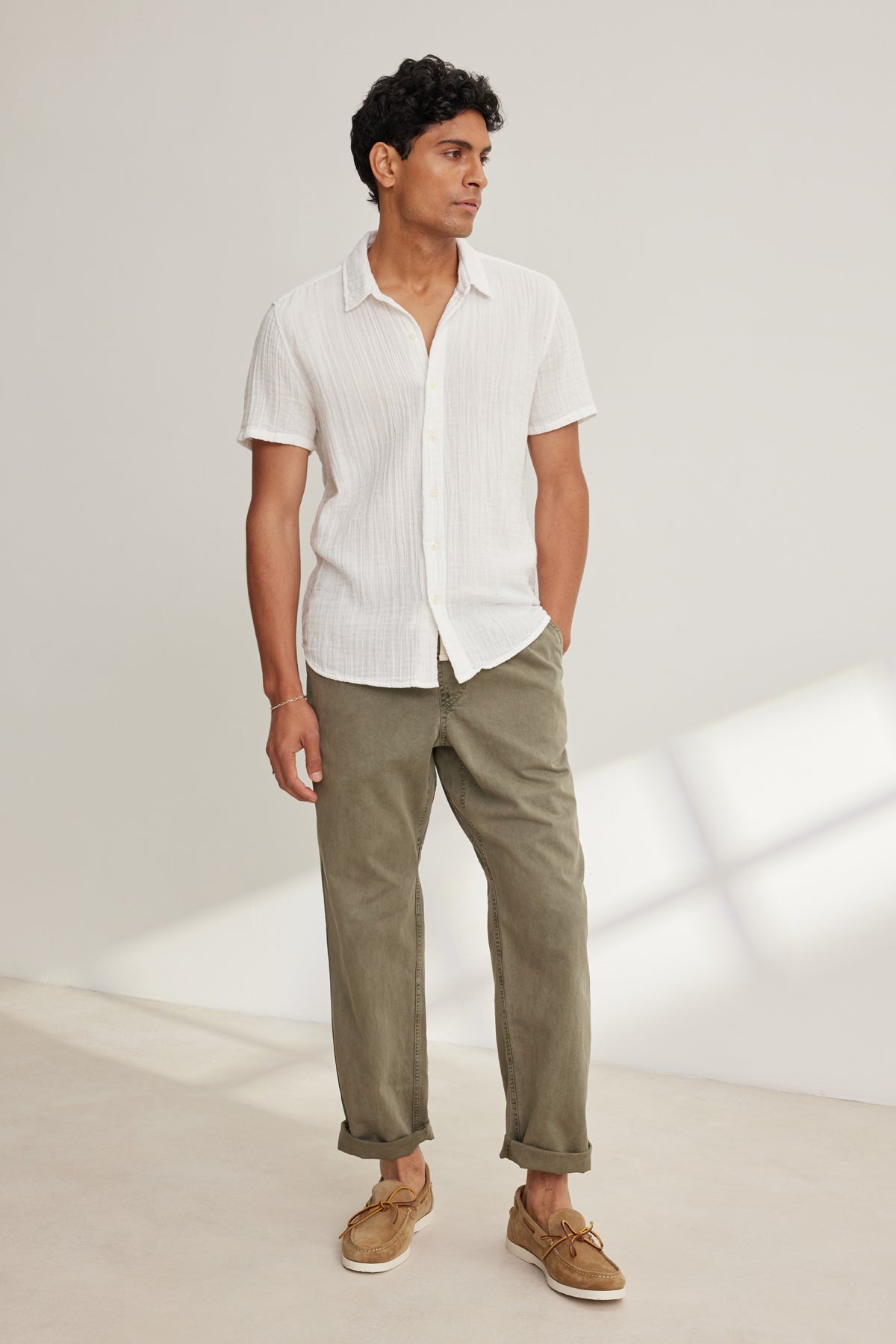 A man stands indoors, hands in pockets, wearing a short-sleeve white button-up shirt, tan boat shoes, and the BRANSON PANT by Velvet by Graham & Spencer—olive green cotton sanded twill with a straight leg design—looking to the side.