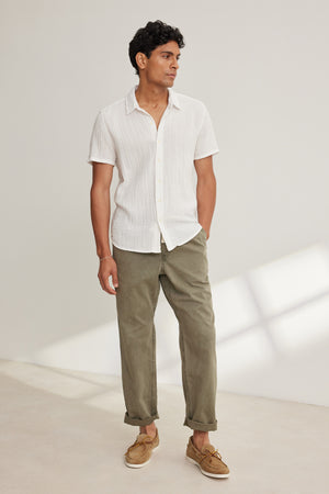 A man stands indoors, hands in pockets, wearing a short-sleeve white button-up shirt, tan boat shoes, and the BRANSON PANT by Velvet by Graham & Spencer—olive green cotton sanded twill with a straight leg design—looking to the side.