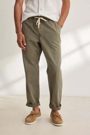 A person wears Velvet by Graham & Spencer's BRANSON PANT in olive green cotton twill with a straight-leg drawstring design, paired with a white short-sleeve shirt and tan boat shoes, standing on a light floor with hands in pockets.
