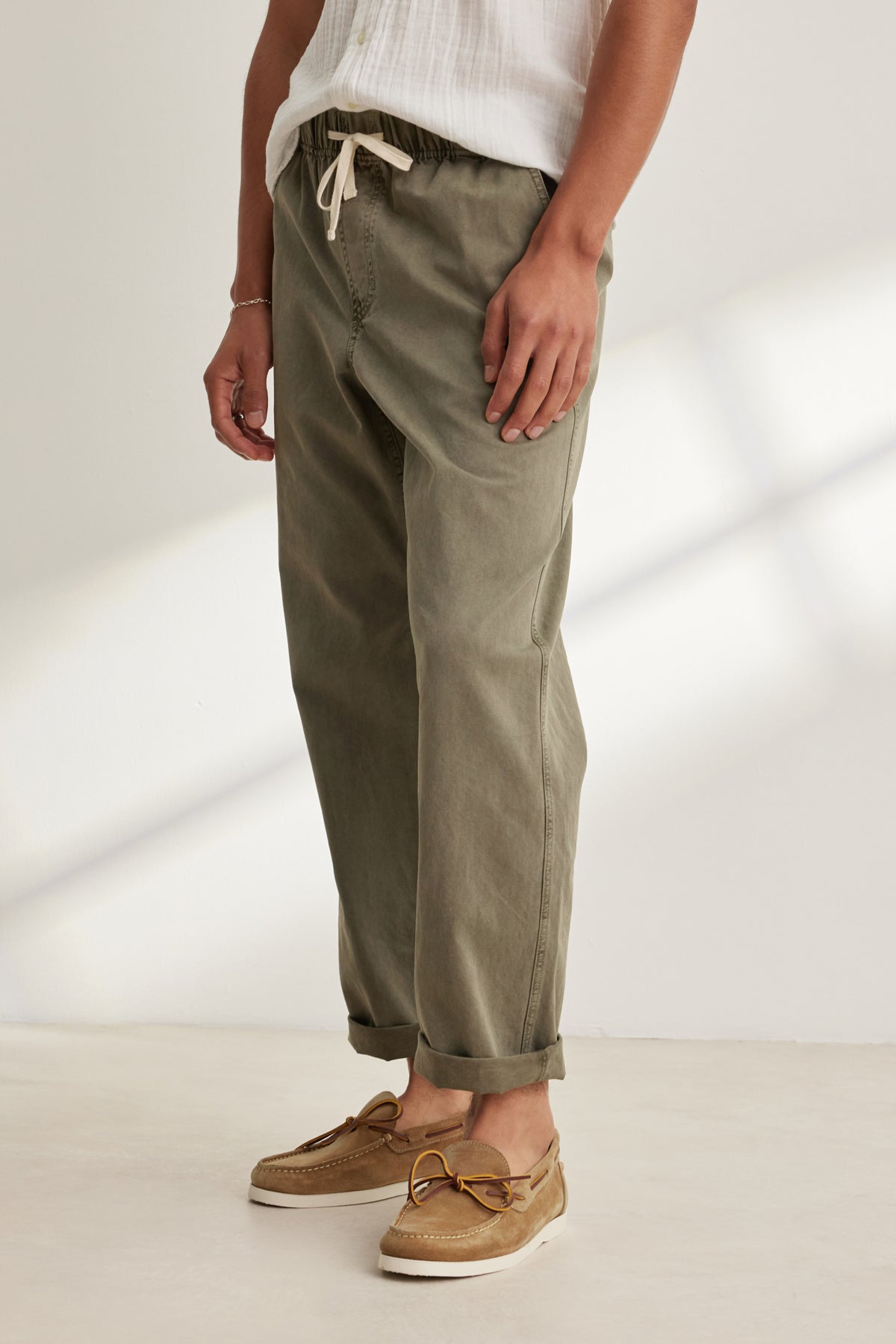 Indoors, a person wears the BRANSON PANT by Velvet by Graham & Spencer in olive green, featuring an elastic drawstring waist, straight leg fit with ankle cuffs, paired with a white shirt and tan boat shoes.