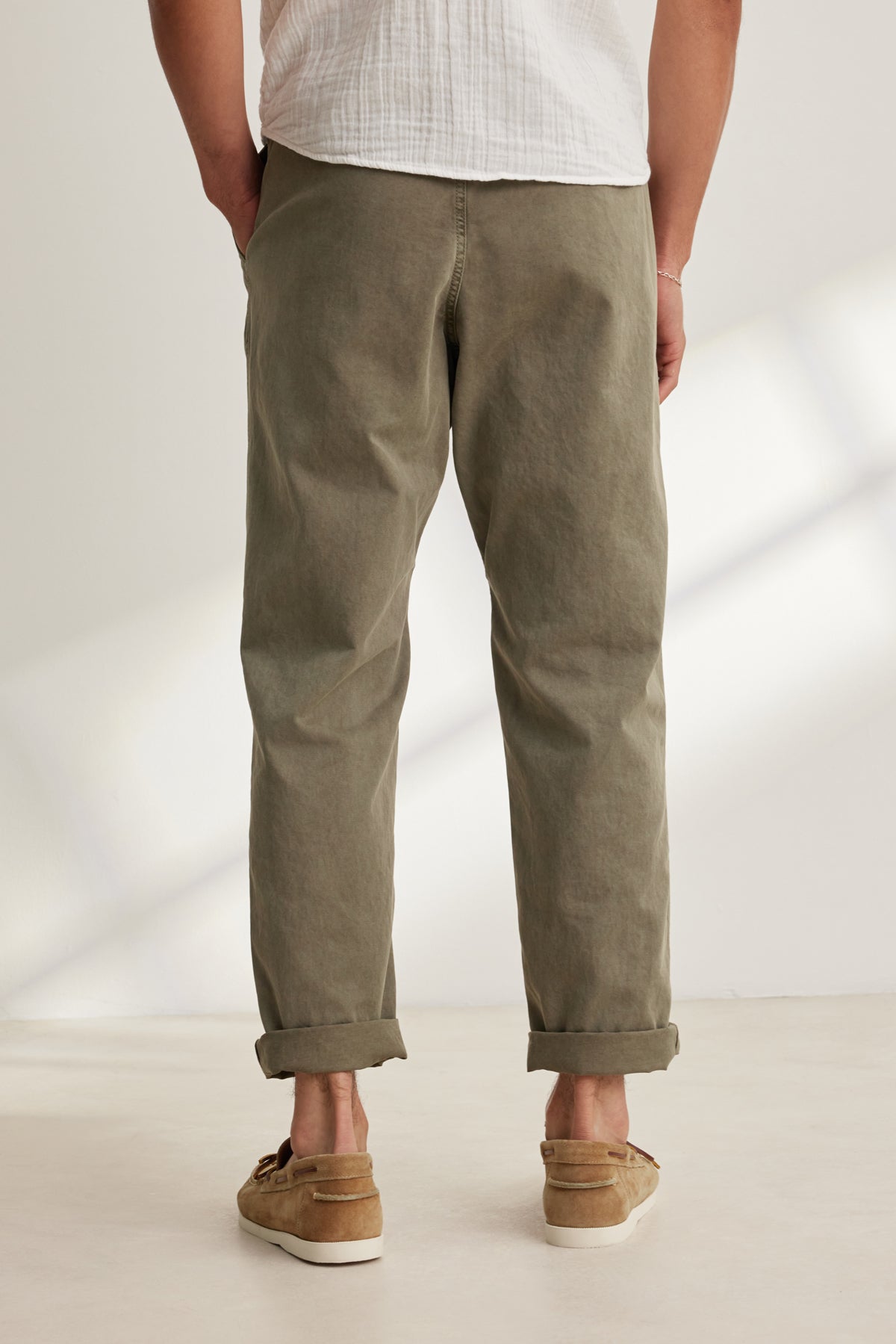A person stands with their back to the camera, wearing the Velvet by Graham & Spencer BRANSON PANT in olive green cotton sanded twill with rolled cuffs, paired with a white shirt and brown loafers.