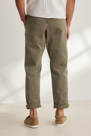 A person stands with their back to the camera, wearing the Velvet by Graham & Spencer BRANSON PANT in olive green cotton sanded twill with rolled cuffs, paired with a white shirt and brown loafers.