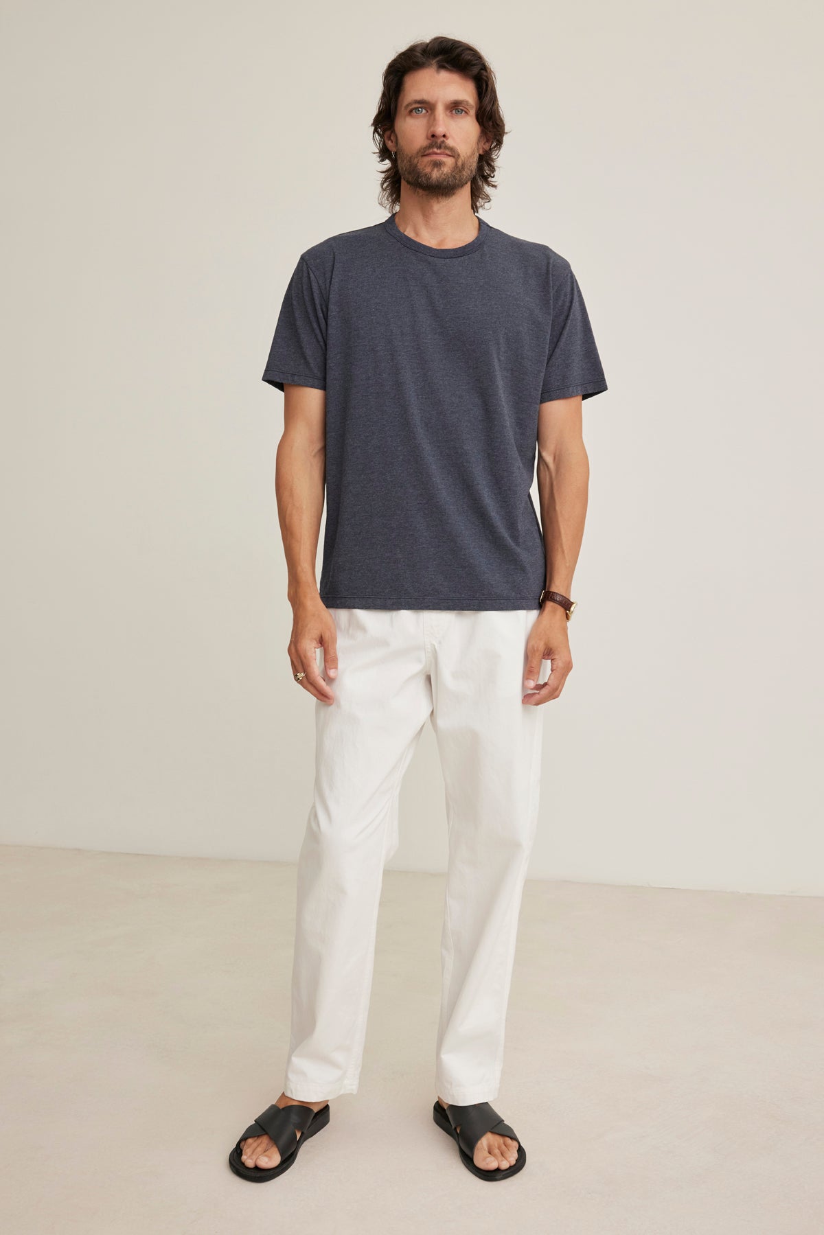 A man stands indoors against a plain light background, wearing the BRANSON PANT by Velvet by Graham & Spencer—white pants with an elastic drawstring waist—paired with a dark gray t-shirt and black sandals.