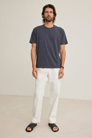 A man stands indoors against a plain light background, wearing the BRANSON PANT by Velvet by Graham & Spencer—white pants with an elastic drawstring waist—paired with a dark gray t-shirt and black sandals.