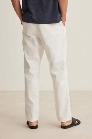 A person stands with hands in pockets, facing away from the camera, wearing a dark shirt and Velvet by Graham & Spencer BRANSON PANT in white cotton sanded twill with a straight leg, paired with black sandals against a plain background.