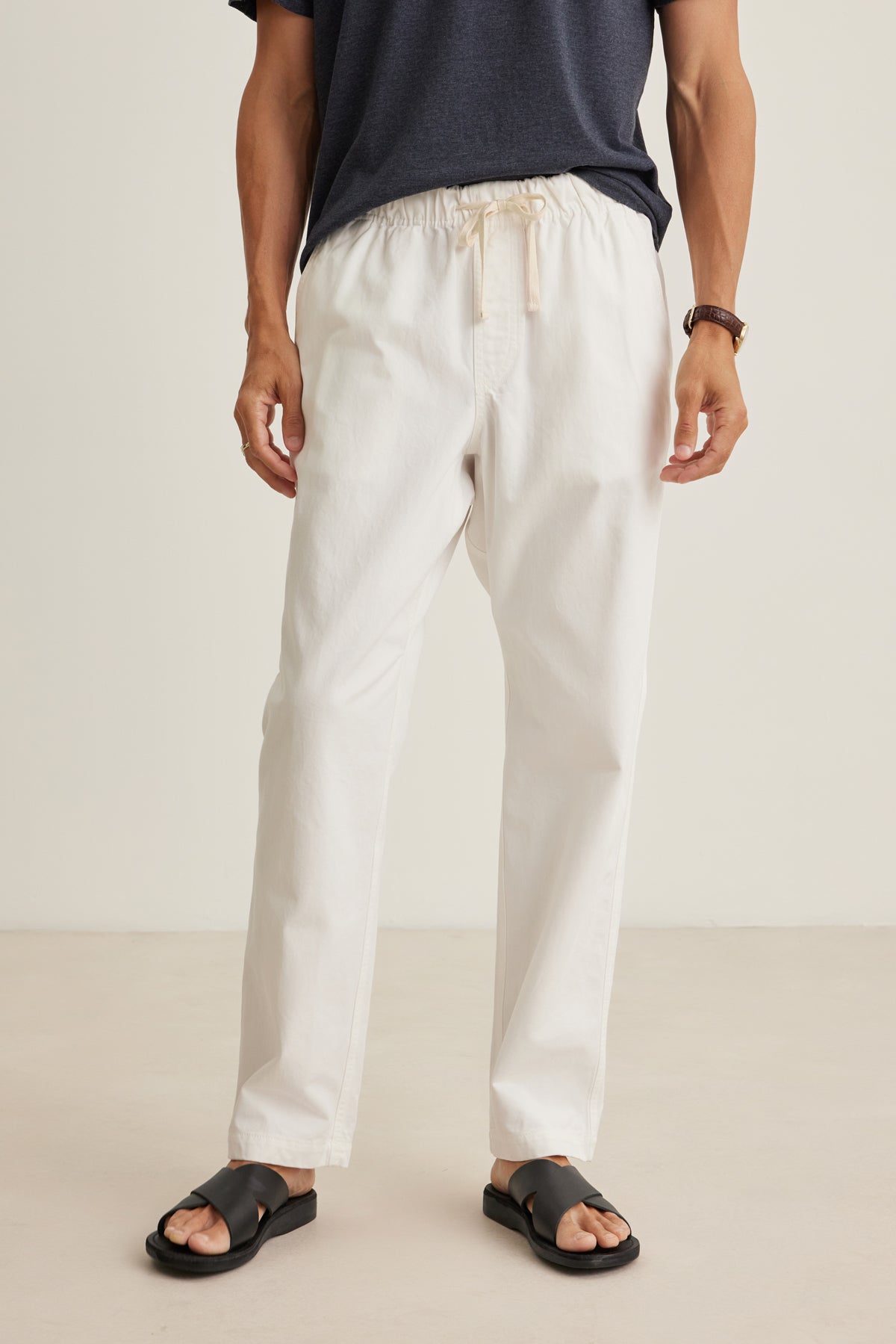 A person wearing a dark t-shirt, Velvet by Graham & Spencer BRANSON PANT in off-white sanded cotton twill with an elastic drawstring waist, black sandals, and a watch stands against a plain light background.