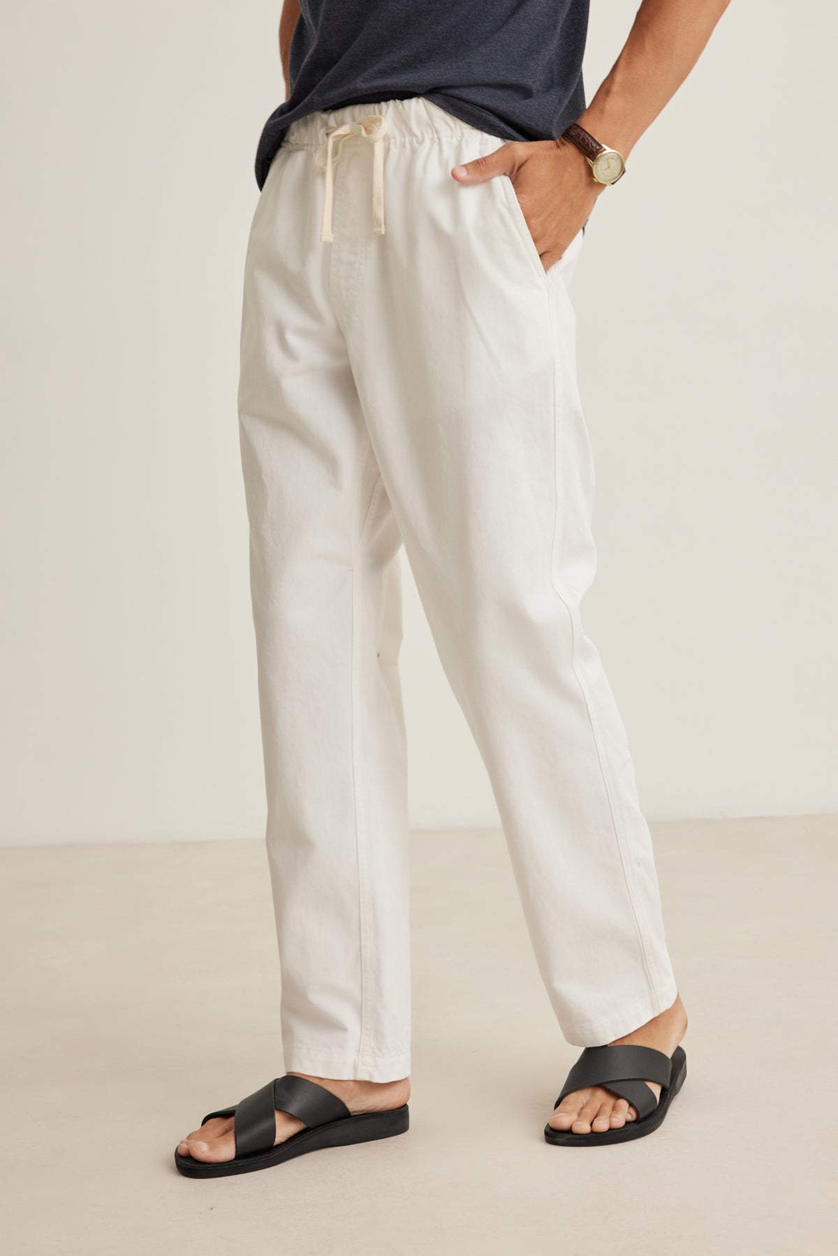 Wearing Velvet by Graham & Spencer’s BRANSON PANT, this person pairs the off-white cotton drawstring pants with a dark shirt, black sandals, and a watch, standing with one hand in their pocket against a plain background.