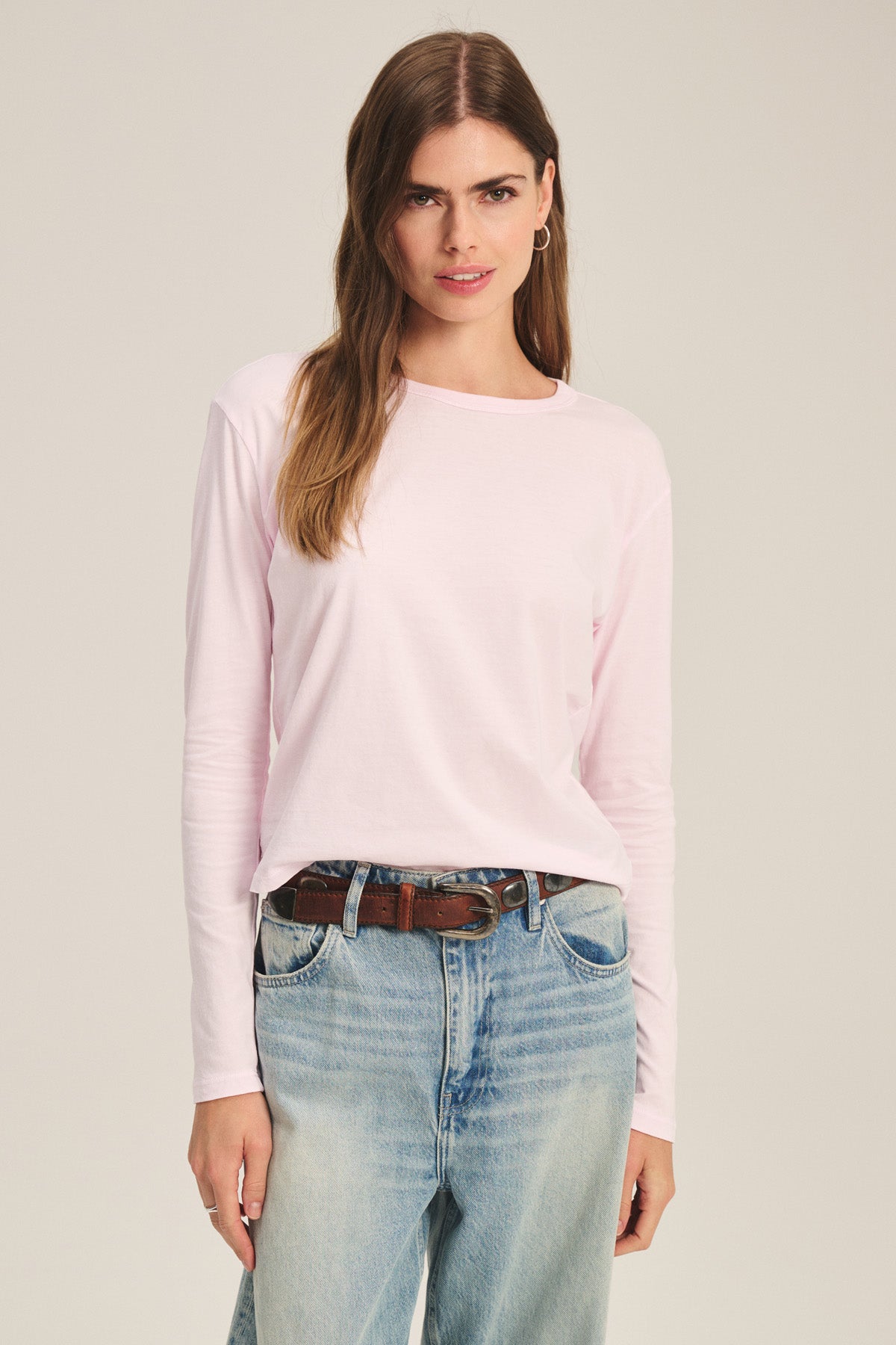 A woman wears the SCOUT TEE by Velvet by Graham & Spencer in light pink, paired with blue jeans and a brown belt, standing facing forward against a plain background.-41436019949761