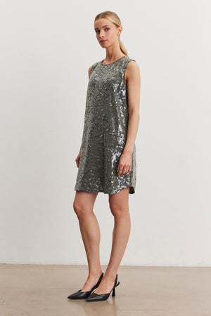 A person stands against a plain white background, wearing the KATHERINE SEQUIN DRESS by Velvet by Graham & Spencer and black heels.