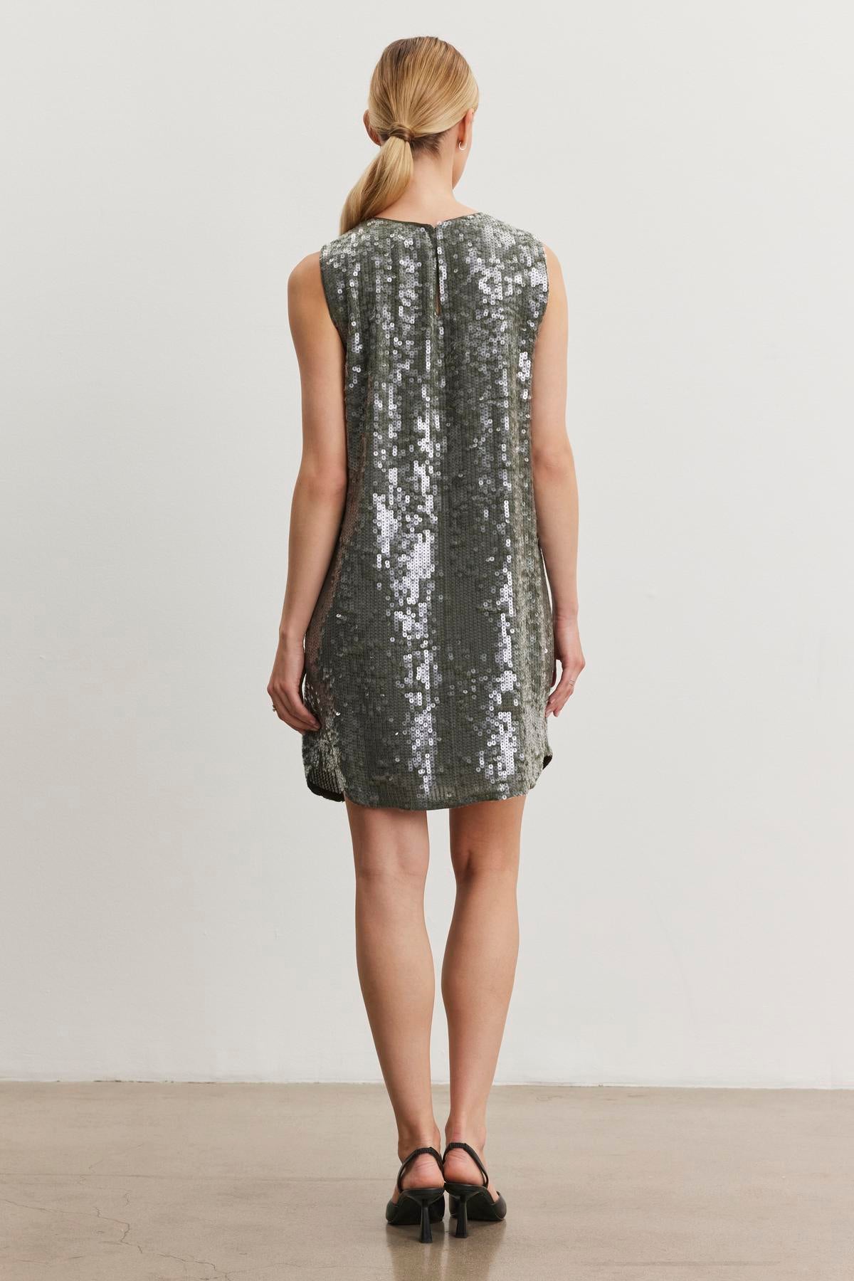 A person in the KATHERINE SEQUIN DRESS from Velvet by Graham & Spencer stands facing a light wall, with hair tied back.-38246520750273