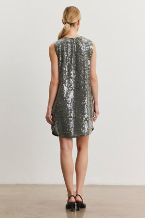 A person in the KATHERINE SEQUIN DRESS from Velvet by Graham & Spencer stands facing a light wall, with hair tied back.
