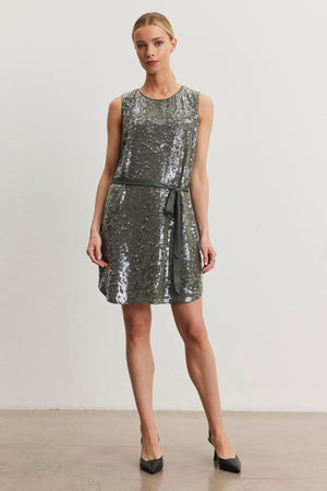 Wearing the KATHERINE SEQUIN DRESS by Velvet by Graham & Spencer, a person poses against a plain backdrop in a sleeveless, knee-length silver sequin dress with a ribbon belt paired with black pointed heels.
