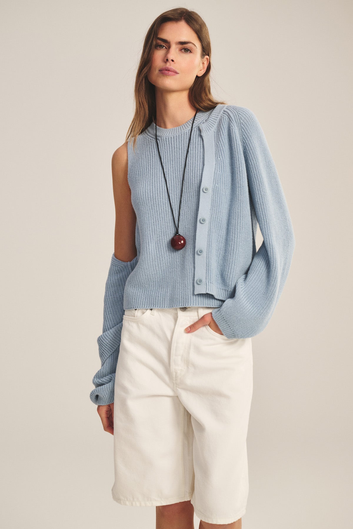 A woman wears the MIRAE CARDIGAN by Velvet by Graham & Spencer over a light blue asymmetrical knit top, paired with white knee-length shorts and a long pendant necklace, standing with one hand in her pocket.-40909904150721