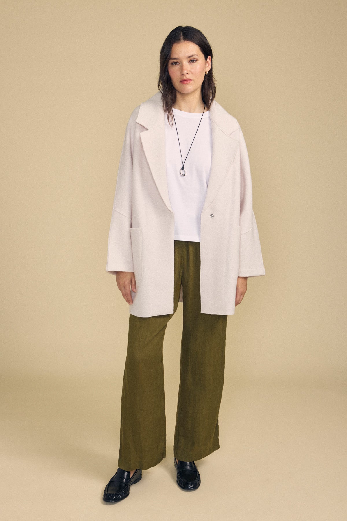 A woman stands against a beige background wearing the ABBY BLAZER by Velvet by Graham & Spencer—an oversized white wool blazer styled with a white shirt, olive green wide-leg pants, black shoes, and a black necklace—perfect for everyday layering.-41860415520961