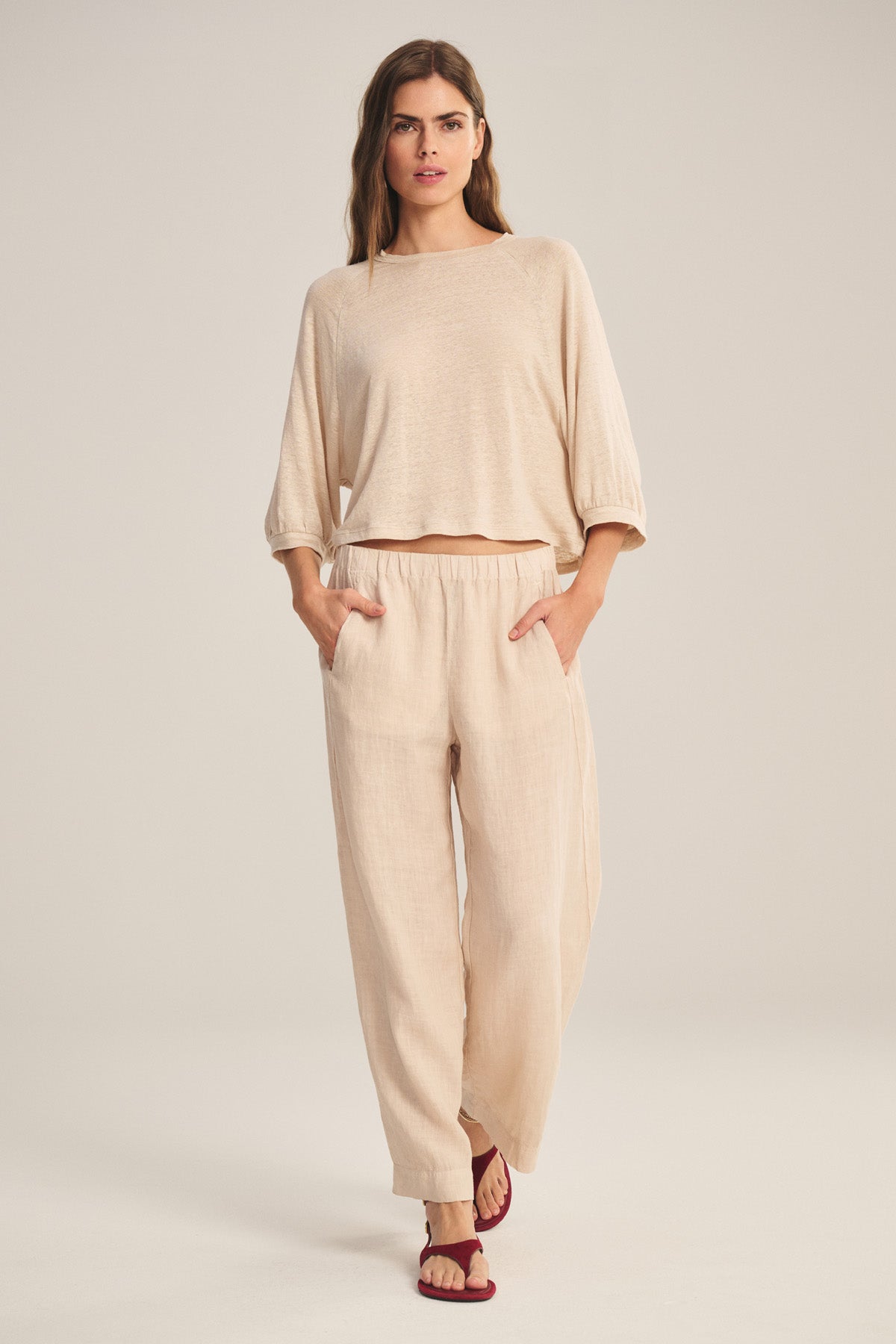 A woman stands facing forward wearing the Velvet by Graham & Spencer LOLA LINEN PANT, a lightweight linen trouser with an elastic waist, paired with a loose beige long-sleeve top and red sandals against a plain background.-40909840646337