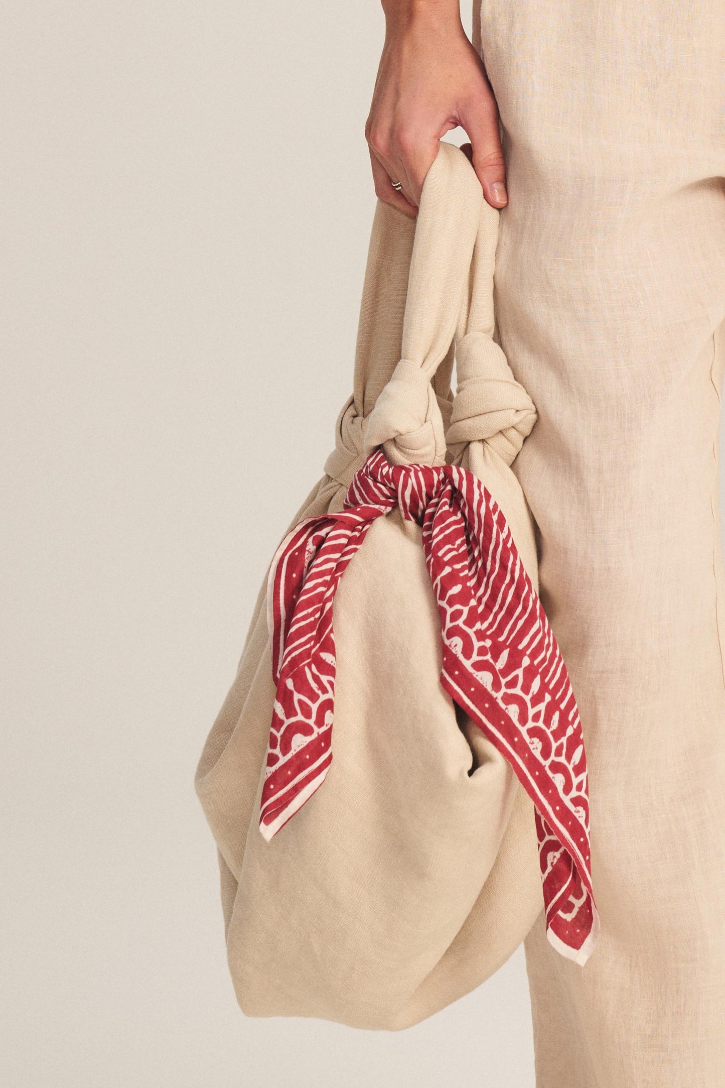 A person holds a beige fabric bag with knotted handles, accessorized with the LISETTE SCARF by Velvet by Graham & Spencer—a red patterned, lightweight cotton scarf—while dressed in beige linen clothing.-40826385694913