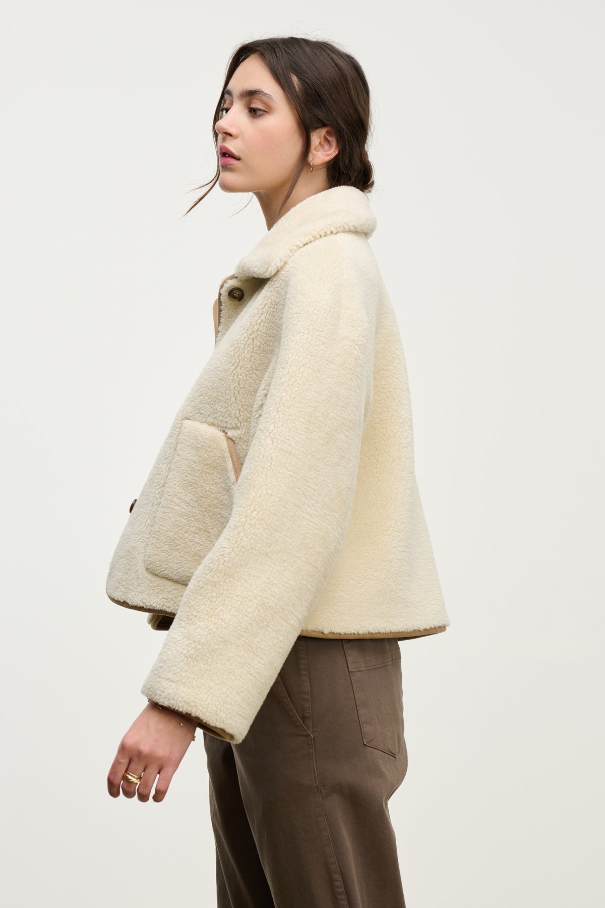 A woman stands sideways against a plain light background wearing the AMARA SHERPA REVERSIBLE JACKET by Velvet by Graham & Spencer in cream, paired with brown pants.-39814804668609