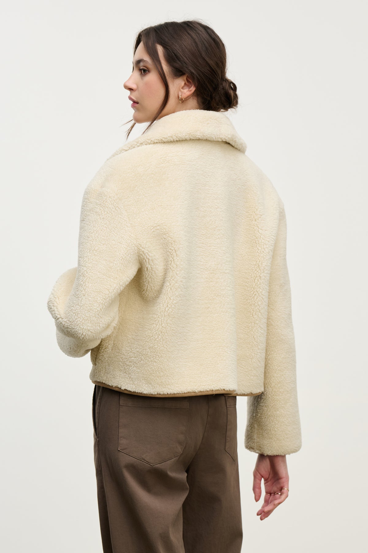 A woman with brown hair in a low bun stands with her back to the camera, wearing the Velvet by Graham & Spencer AMARA SHERPA REVERSIBLE JACKET in beige and brown pants against a plain background.