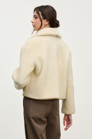 A woman with brown hair in a low bun stands with her back to the camera, wearing the Velvet by Graham & Spencer AMARA SHERPA REVERSIBLE JACKET in beige and brown pants against a plain background.