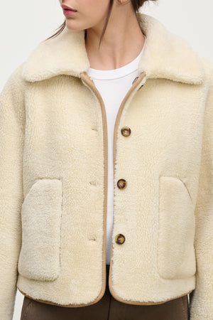 A woman wearing the AMARA SHERPA REVERSIBLE JACKET by Velvet by Graham & Spencer—a cream faux sherpa jacket with large front pockets and brown buttons—over a white shirt and brown pants.