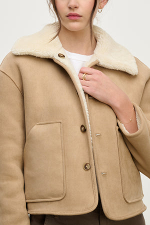 A woman wears the AMARA SHERPA REVERSIBLE JACKET by Velvet by Graham & Spencer—beige faux sherpa with large front pockets, styled over a white shirt. She holds the collar of the jacket with one hand.