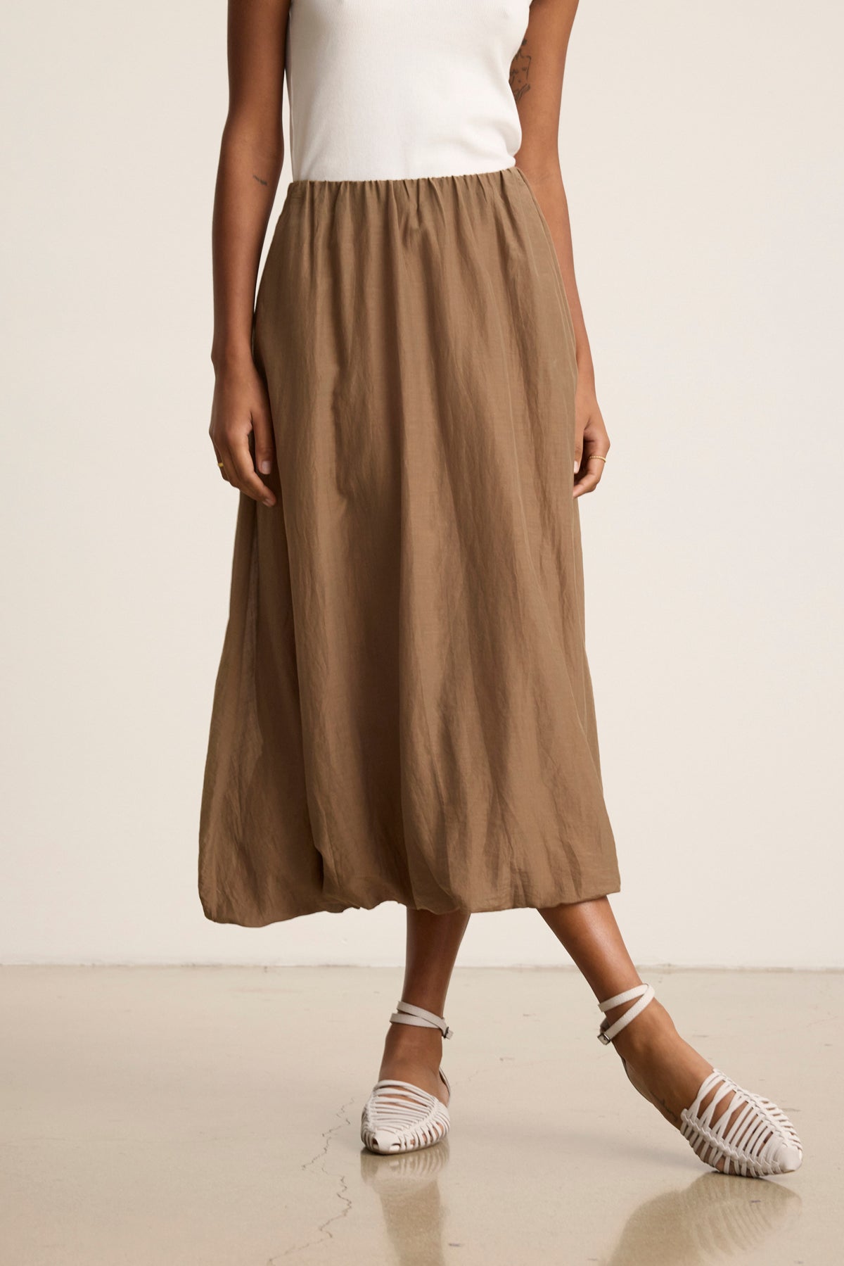 A person wears a white sleeveless top, the INES SILK COTTON VOILE SKIRT by Velvet by Graham & Spencer in tan with an elastic waist, and white woven flat shoes, standing against a plain background.