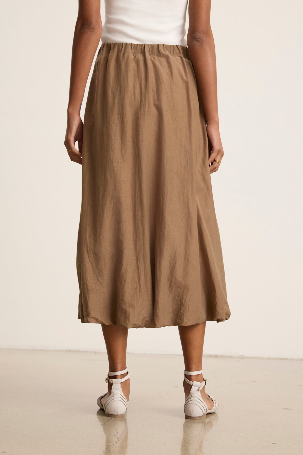 A person is shown from the back against a neutral background, wearing a white top, white sandals, and the INES SILK COTTON VOILE SKIRT by Velvet by Graham & Spencer—a tan midi skirt with an elastic waist.