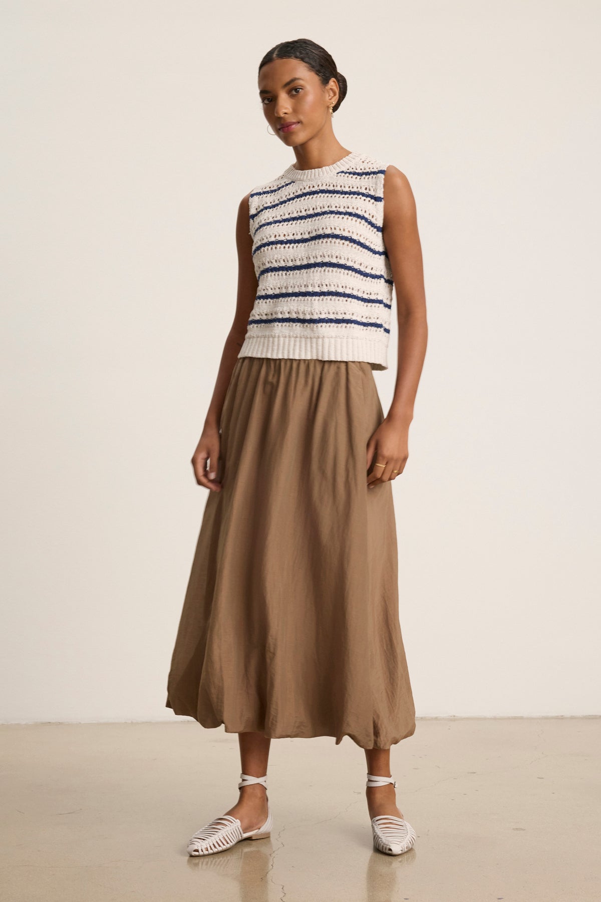 A woman stands indoors against a plain background wearing the INES SILK COTTON VOILE SKIRT by Velvet by Graham & Spencer, paired with a sleeveless striped knit top and white woven flat shoes.-39119890776257