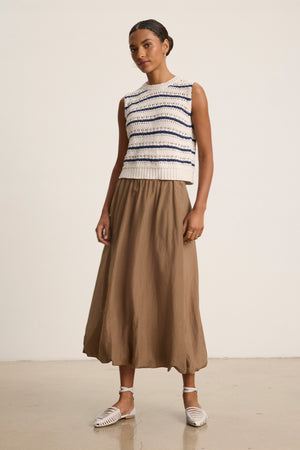 A woman stands indoors against a plain background wearing the INES SILK COTTON VOILE SKIRT by Velvet by Graham & Spencer, paired with a sleeveless striped knit top and white woven flat shoes.
