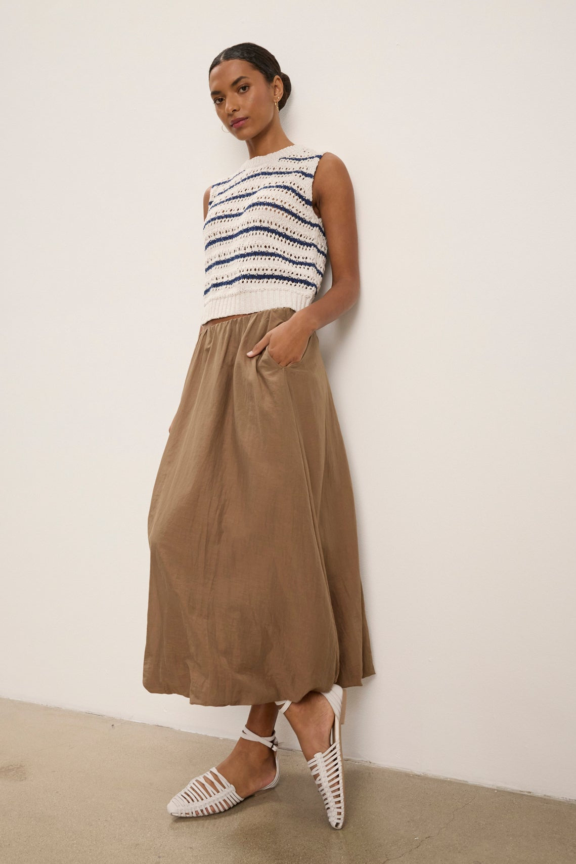 Woman standing against a plain wall, wearing a sleeveless white and navy striped top, the INES SILK COTTON VOILE SKIRT by Velvet by Graham & Spencer—a long tan bubble hem skirt with elastic waist—and white woven flats.