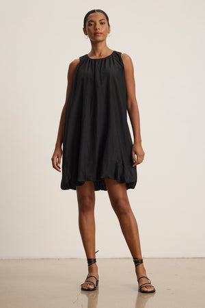 A woman stands against a plain background wearing the Velvet by Graham & Spencer MARGOT SILK COTTON VOILE DRESS, a sleeveless, loose black dress, paired with black strappy sandals.