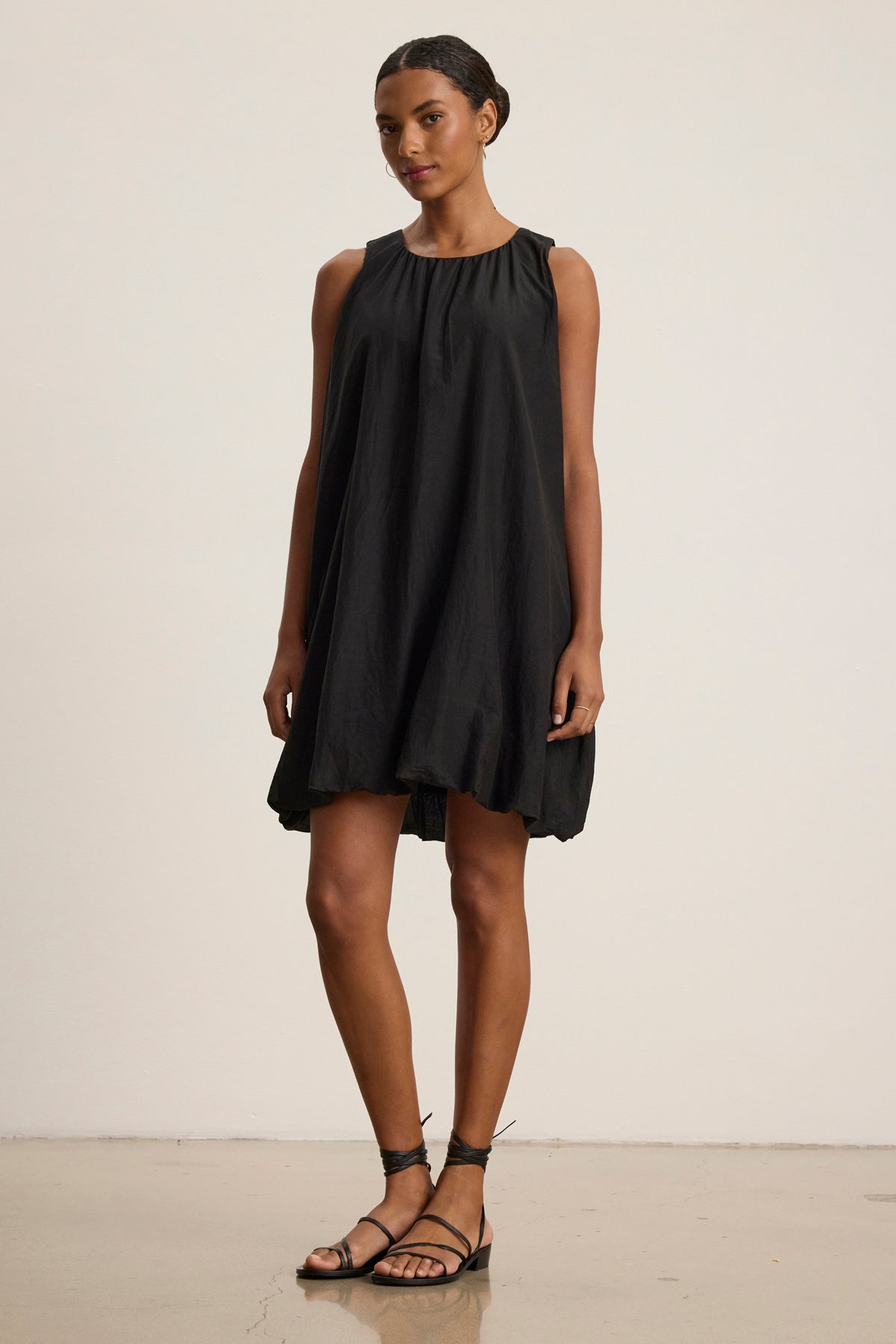A woman poses against a plain background in the MARGOT SILK COTTON VOILE DRESS by Velvet by Graham & Spencer—a sleeveless, modern boxy fit black dress—paired with black strappy sandals.-39111855767745