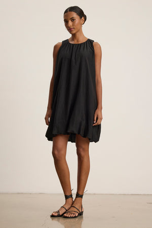 A woman poses against a plain background in the MARGOT SILK COTTON VOILE DRESS by Velvet by Graham & Spencer—a sleeveless, modern boxy fit black dress—paired with black strappy sandals.