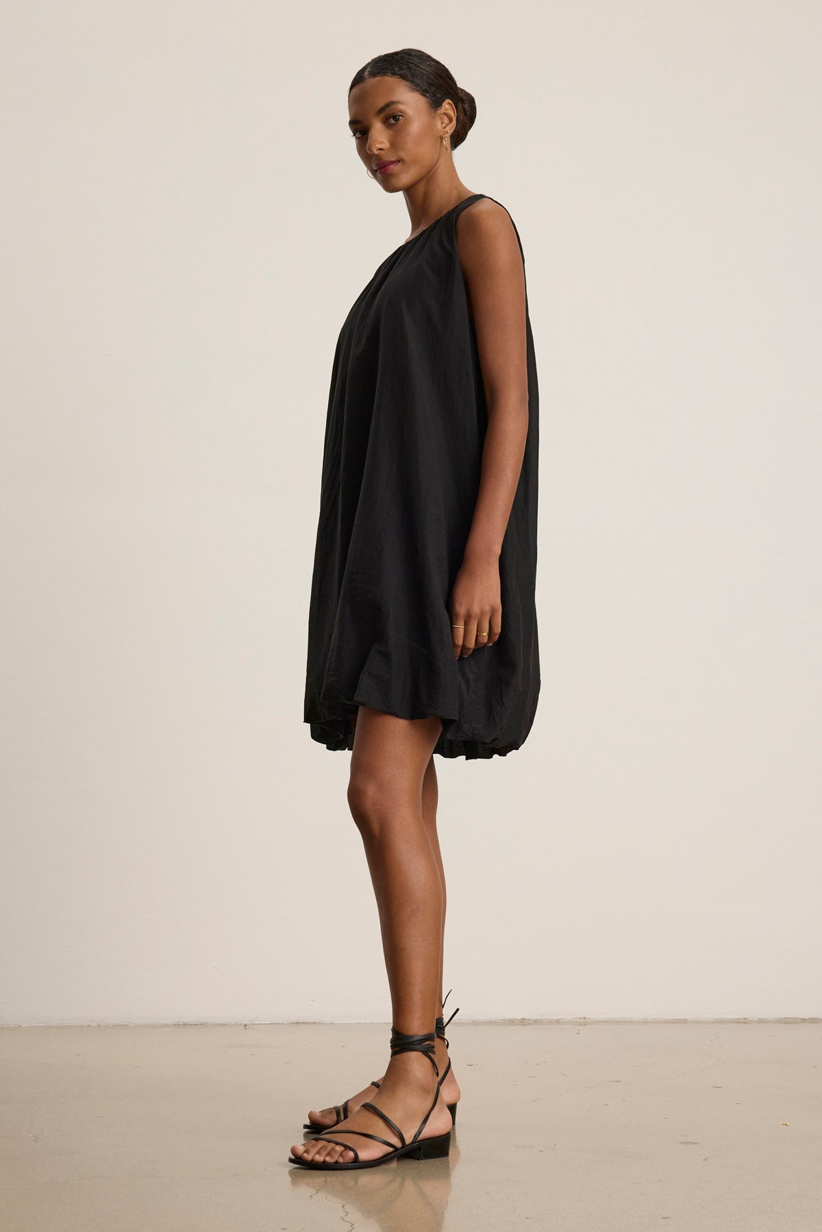 A woman stands sideways in the MARGOT SILK COTTON VOILE DRESS by Velvet by Graham & Spencer, a loose-fitting black sleeveless dress, paired with black strappy sandals against a plain light background.-39111855571137