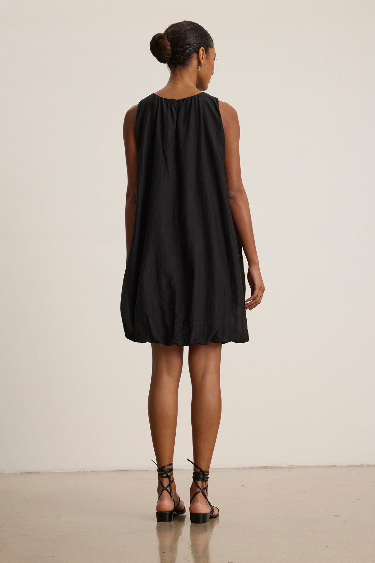 A woman faces away, wearing the MARGOT SILK COTTON VOILE DRESS by Velvet by Graham & Spencer—a sleeveless black dress with a detachable belt—paired with black strappy sandals, on a plain background.-39111855243457