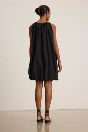 A woman faces away, wearing the MARGOT SILK COTTON VOILE DRESS by Velvet by Graham & Spencer—a sleeveless black dress with a detachable belt—paired with black strappy sandals, on a plain background.