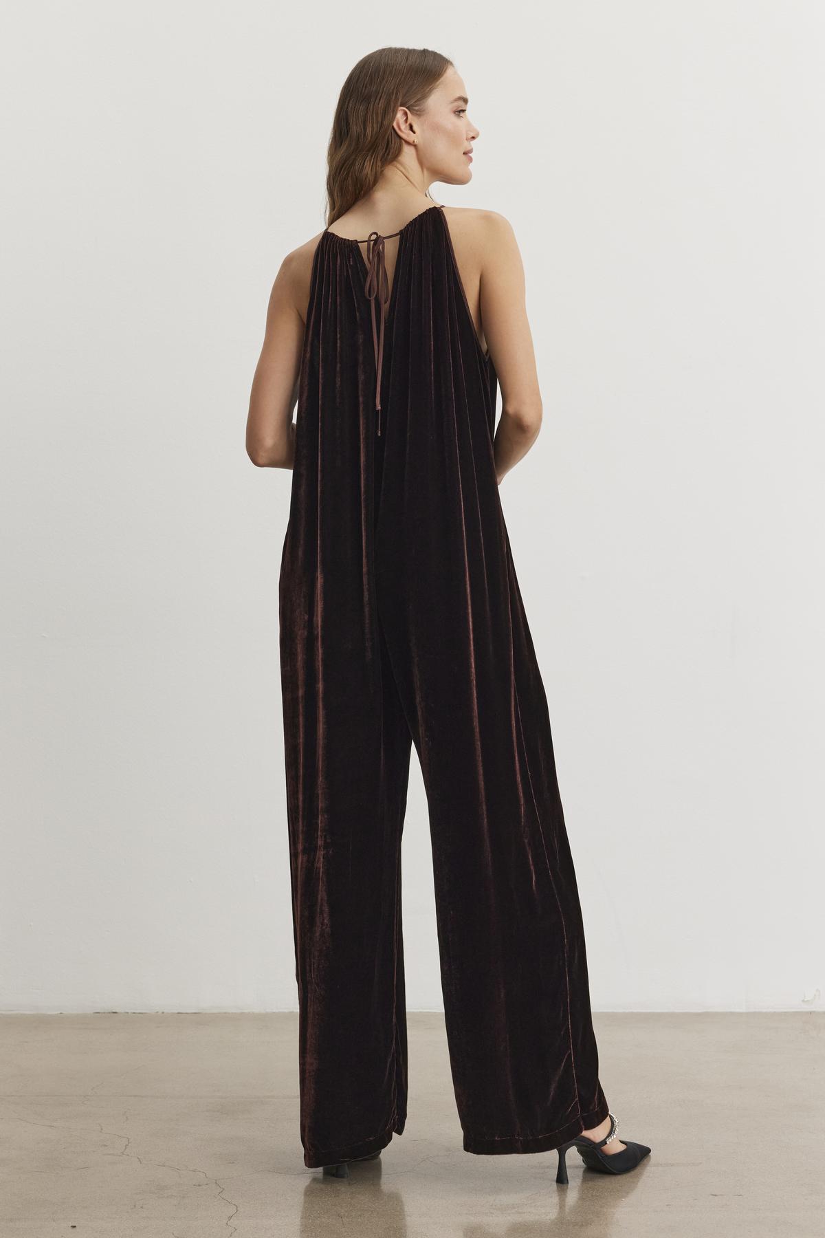 A woman stands with her back to the camera in a neutral room, wearing the Cienna Silk Velvet Jumpsuit by Velvet by Graham & Spencer, paired with black heels.-38246397345985